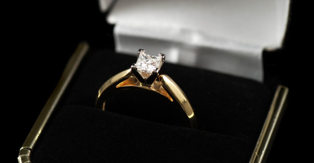 Close-up of a gold engagement ring with a square-cut diamond solitaire, displayed in a black velvet ring box with a white satin interior.