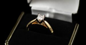 Close-up of a gold engagement ring with a square-cut diamond solitaire, displayed in a black velvet ring box with a white satin interior.