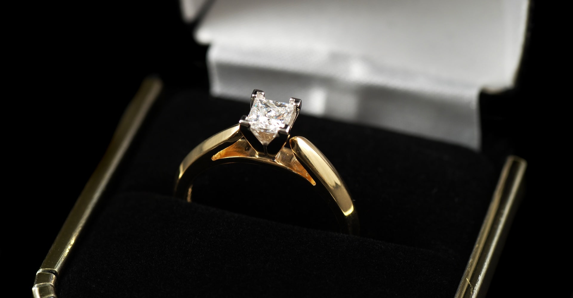 Close-up of a gold engagement ring with a square-cut diamond solitaire, displayed in a black velvet ring box with a white satin interior.