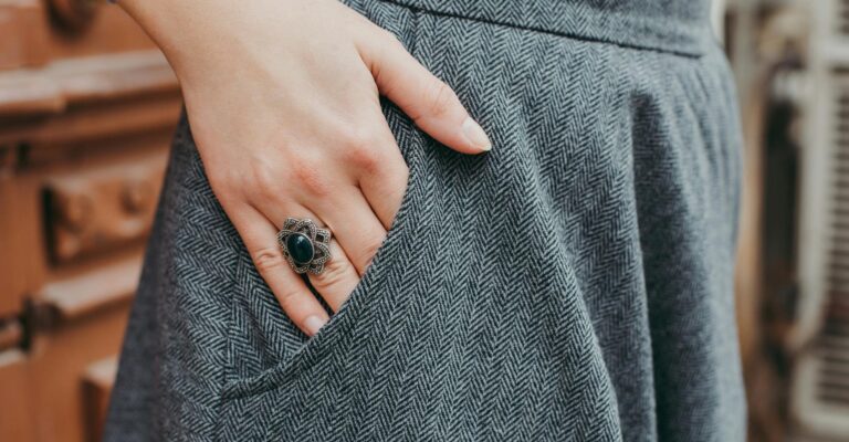 A close-up of a person’s hand resting in the pocket of a gray herringbone-patterned skirt, showcasing a large, vintage-style ring with a black oval stone and ornate silver setting.